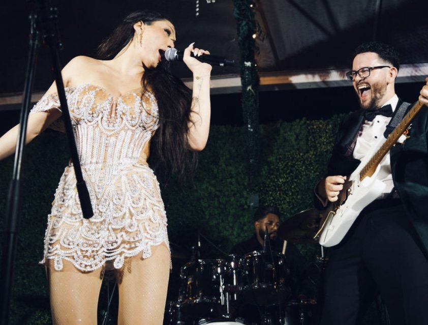 Singer in a sparkling white outfit performs with a guitarist and drummer onstage.