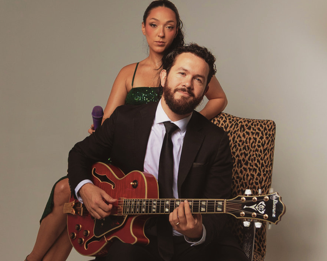 A bearded man in a black suit plays a red hollow-body electric guitar while a woman in a green sequined dress holds a microphone behind him.