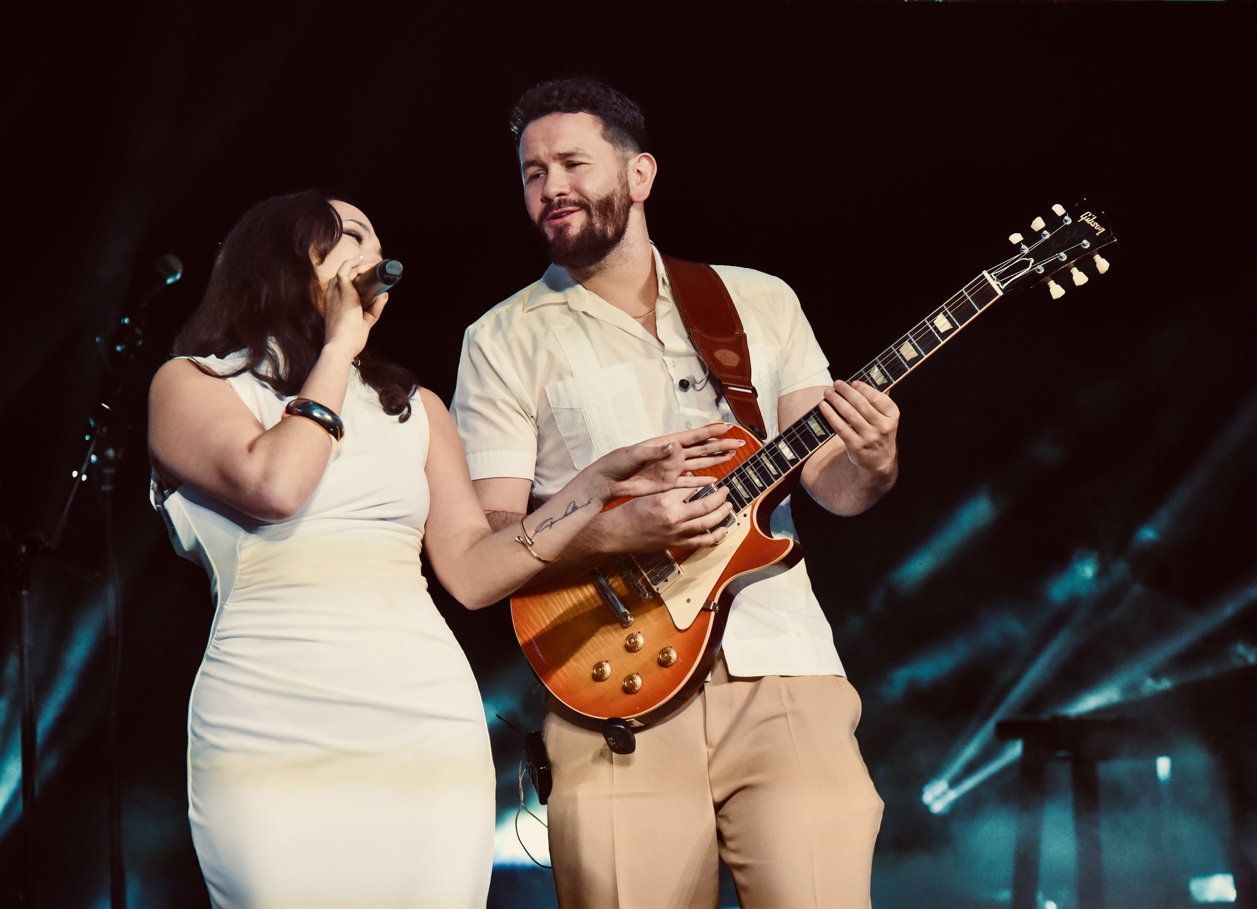 Female singer performing beside a male guitarist playing an electric guitar on a dimly lit concert stage