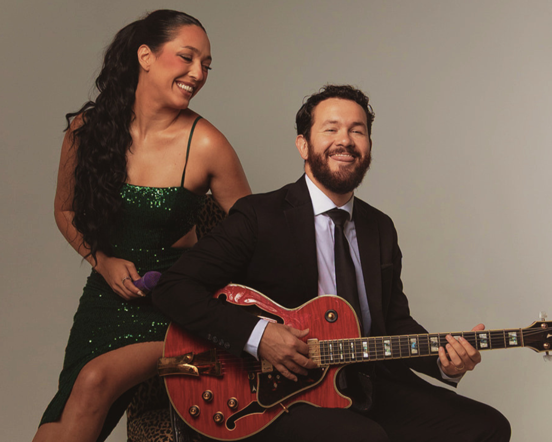 Smiling woman in a green sequined dress stands behind a bearded man in a black suit playing a red electric guitar.