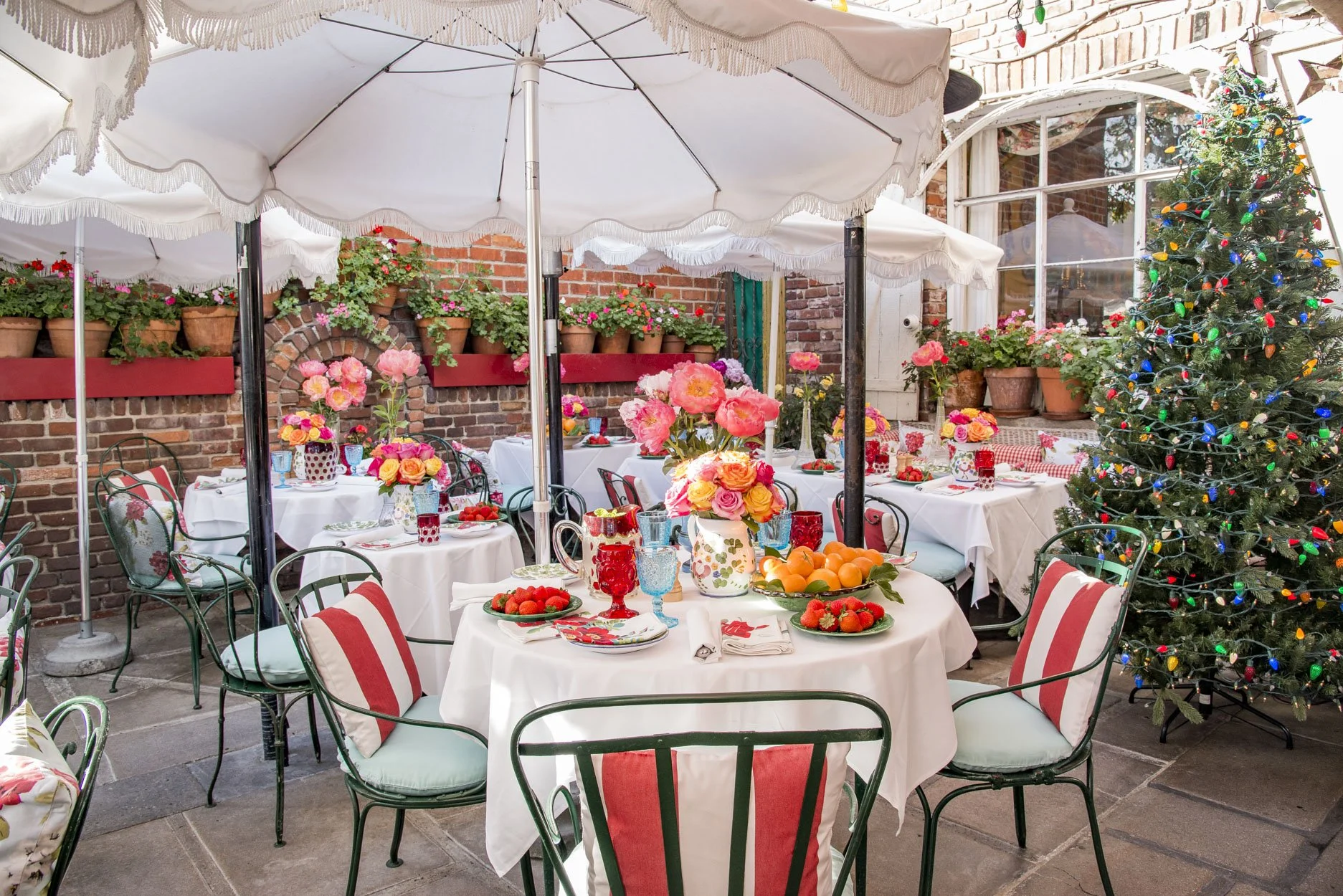 Festive outdoor restaurant patio with white umbrellas, flower-filled tables, and a Christmas tree decorated with colorful lights.