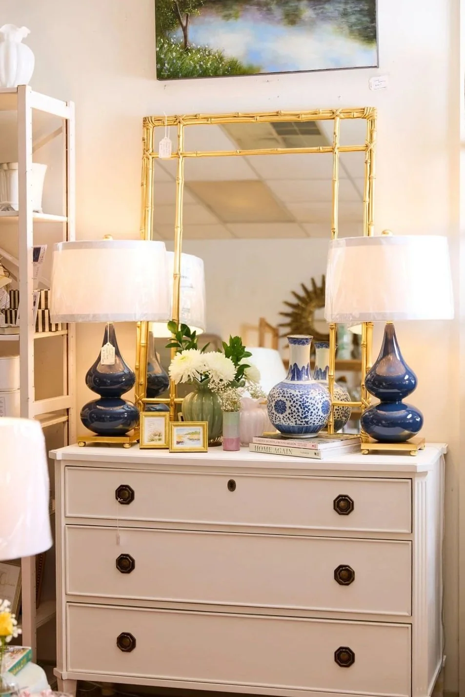 White three-drawer dresser styled with a gold bamboo mirror, blue table lamps, blue-and-white vase, flowers, framed art, and books.