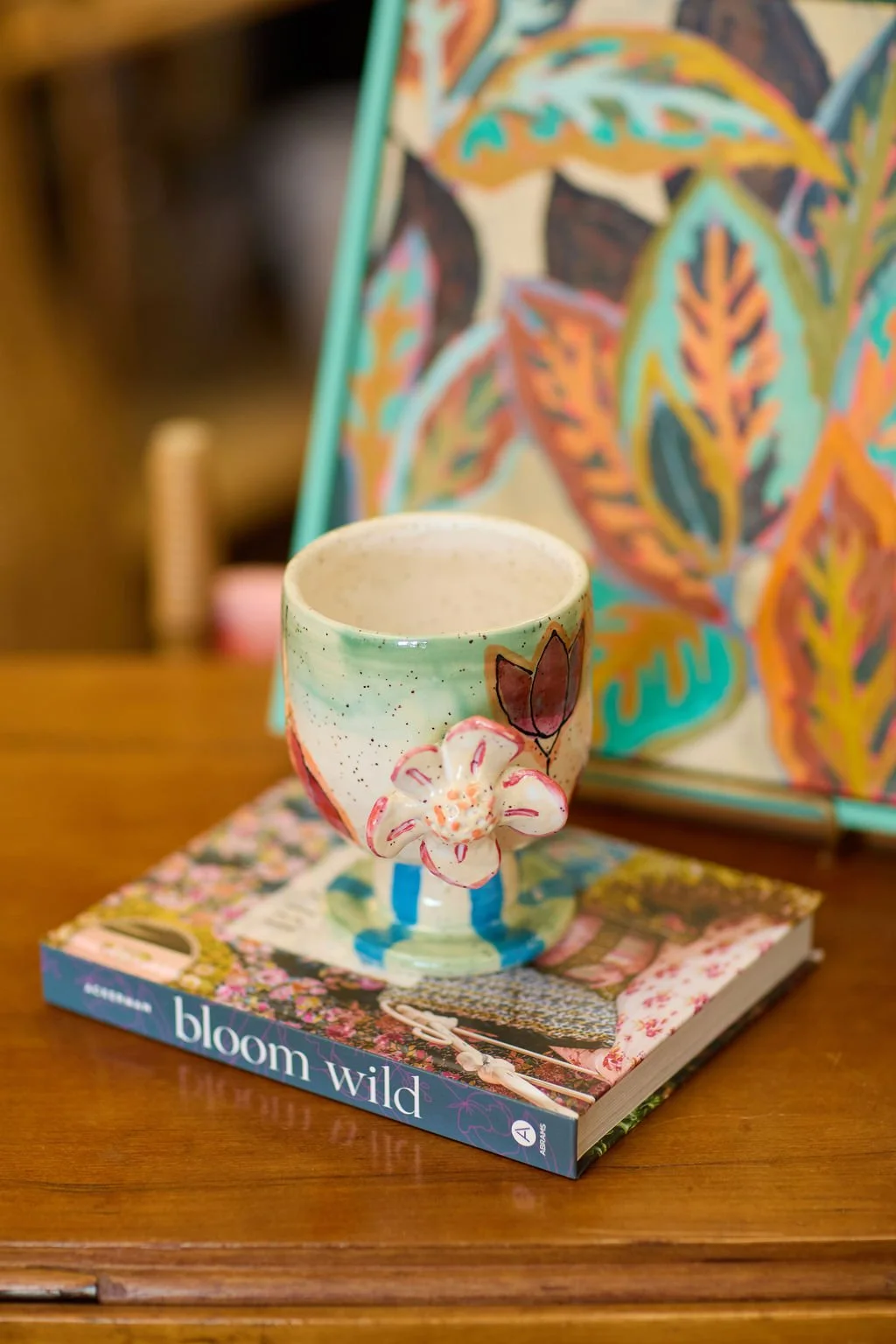 Colorful floral ceramic mug displayed on the book “Bloom Wild” beside a vibrant botanical artwork.