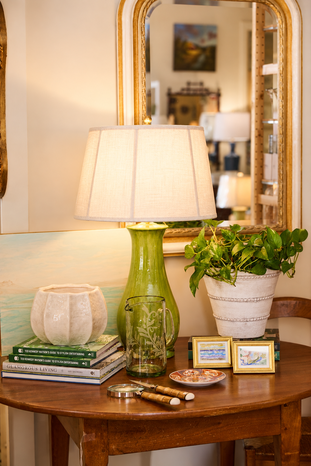 Eclectic entryway vignette with green ceramic table lamp, potted plant, framed artwork, decorative glassware, and books on a rustic wooden console table