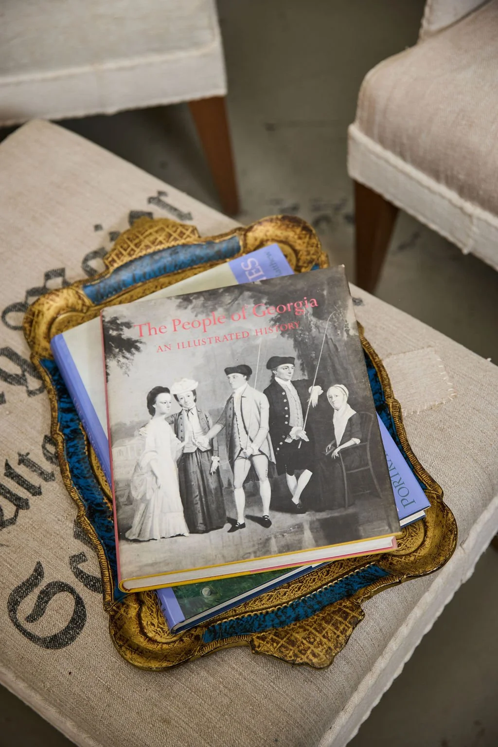 Stack of books, including “The People of Georgia: An Illustrated History,” resting on an ornate gold-and-blue tray atop a beige upholstered chair.