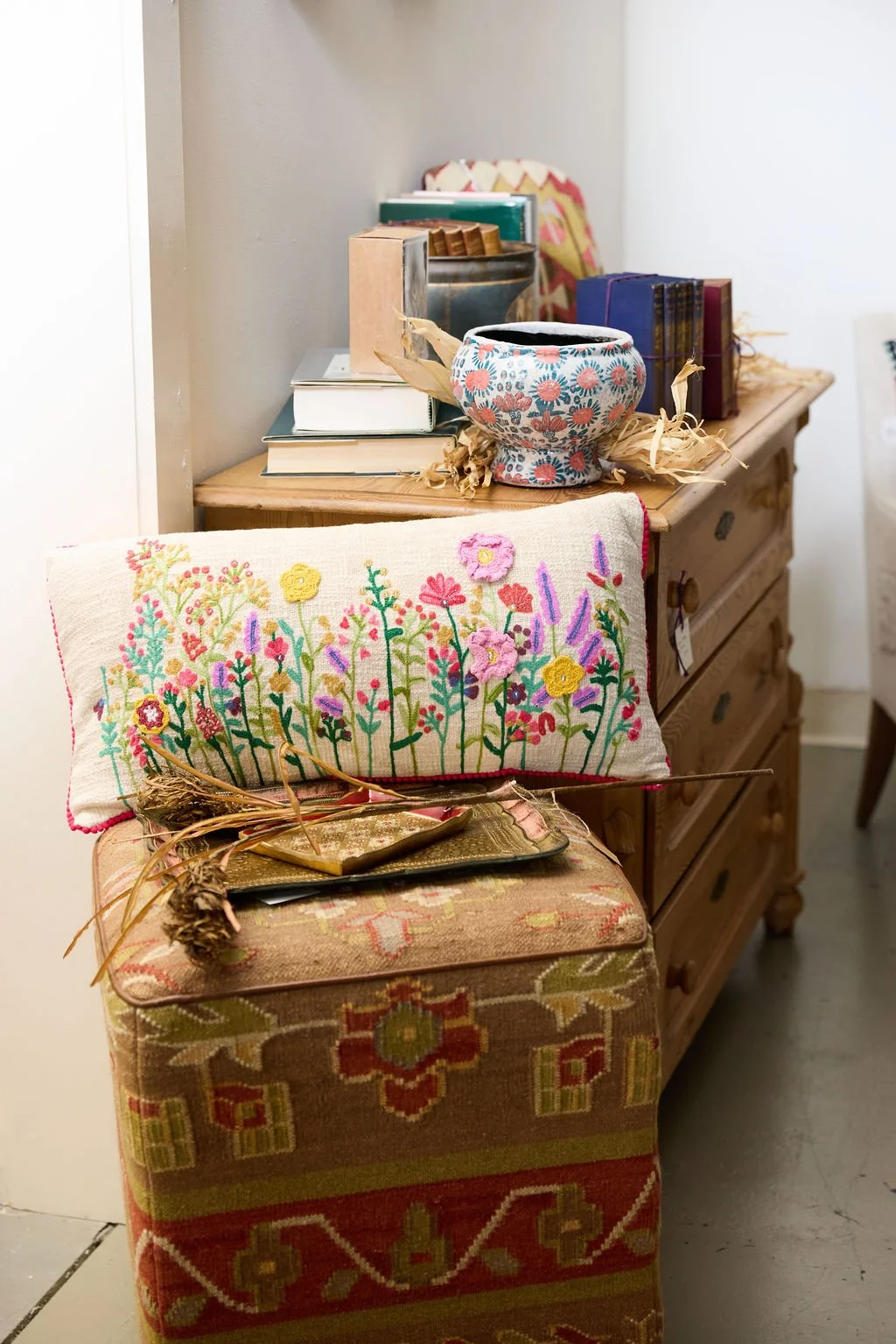 Colorful embroidered floral cushion and patterned ottoman beside a wooden dresser with books and a decorative floral bowl