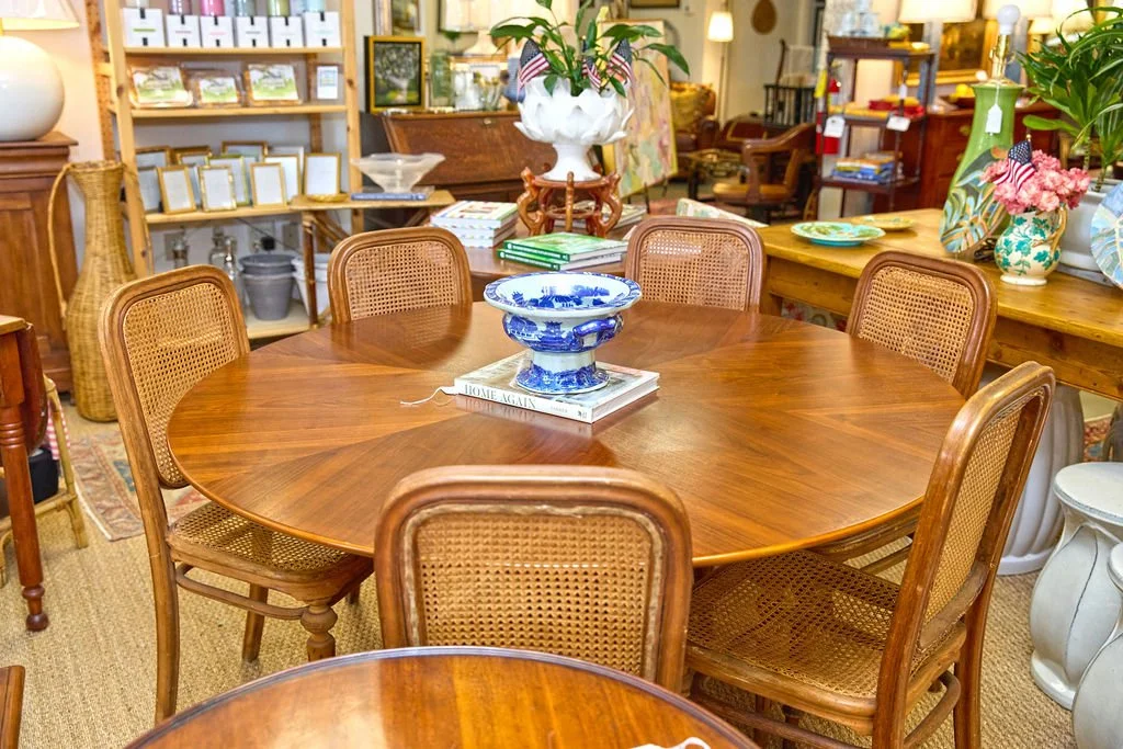 Antique furniture store showroom with a round wooden dining table, cane-back chairs, blue-and-white centerpiece bowl, and decorative furnishings