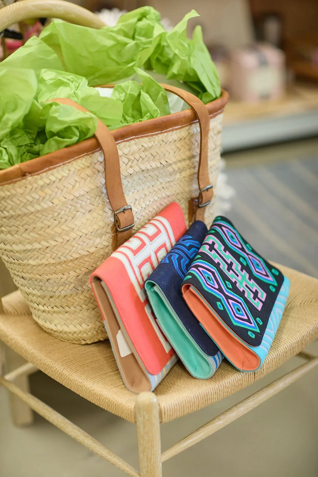 Woven straw tote bag filled with green tissue paper beside colorful rolled textiles on a wicker chair