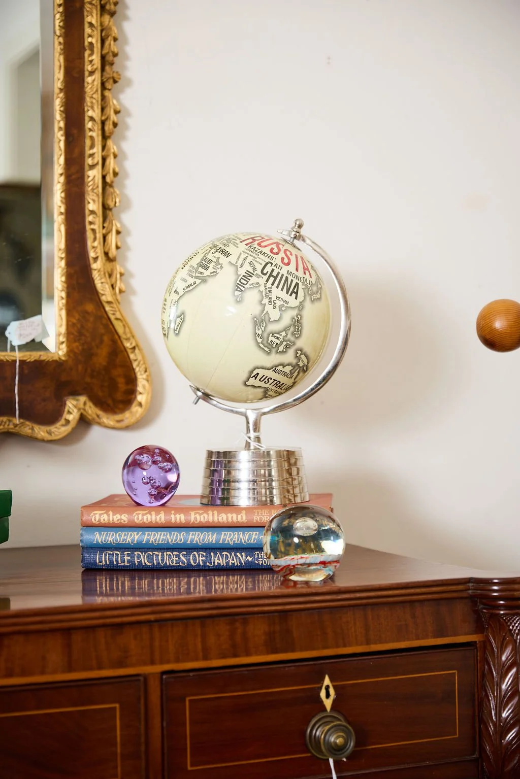Vintage globe displayed on stacked books atop an antique wooden cabinet beside an ornate gold mirror and decorative glass spheres