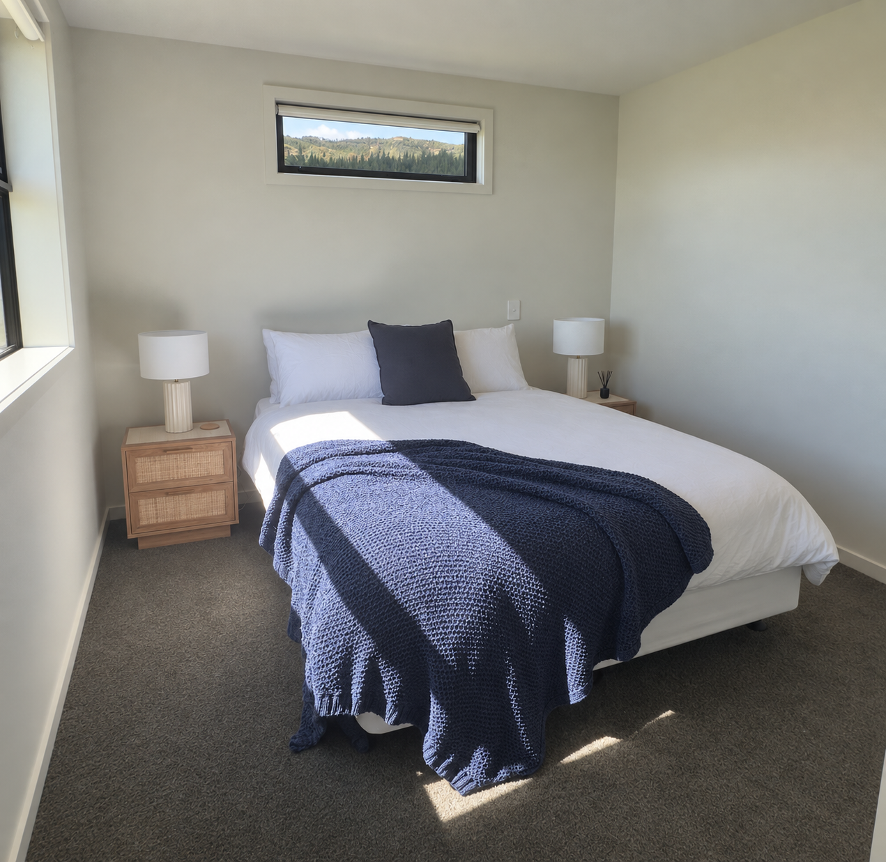 Bright minimalist bedroom with a white bed, navy throw blanket, bedside tables, lamps, and mountain views through the windows