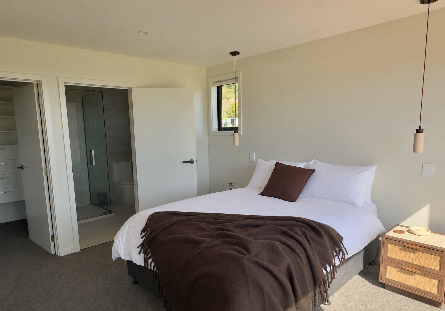 Bright modern bedroom with a white bed, brown throw blanket, pendant lights, ensuite bathroom, and wooden nightstand