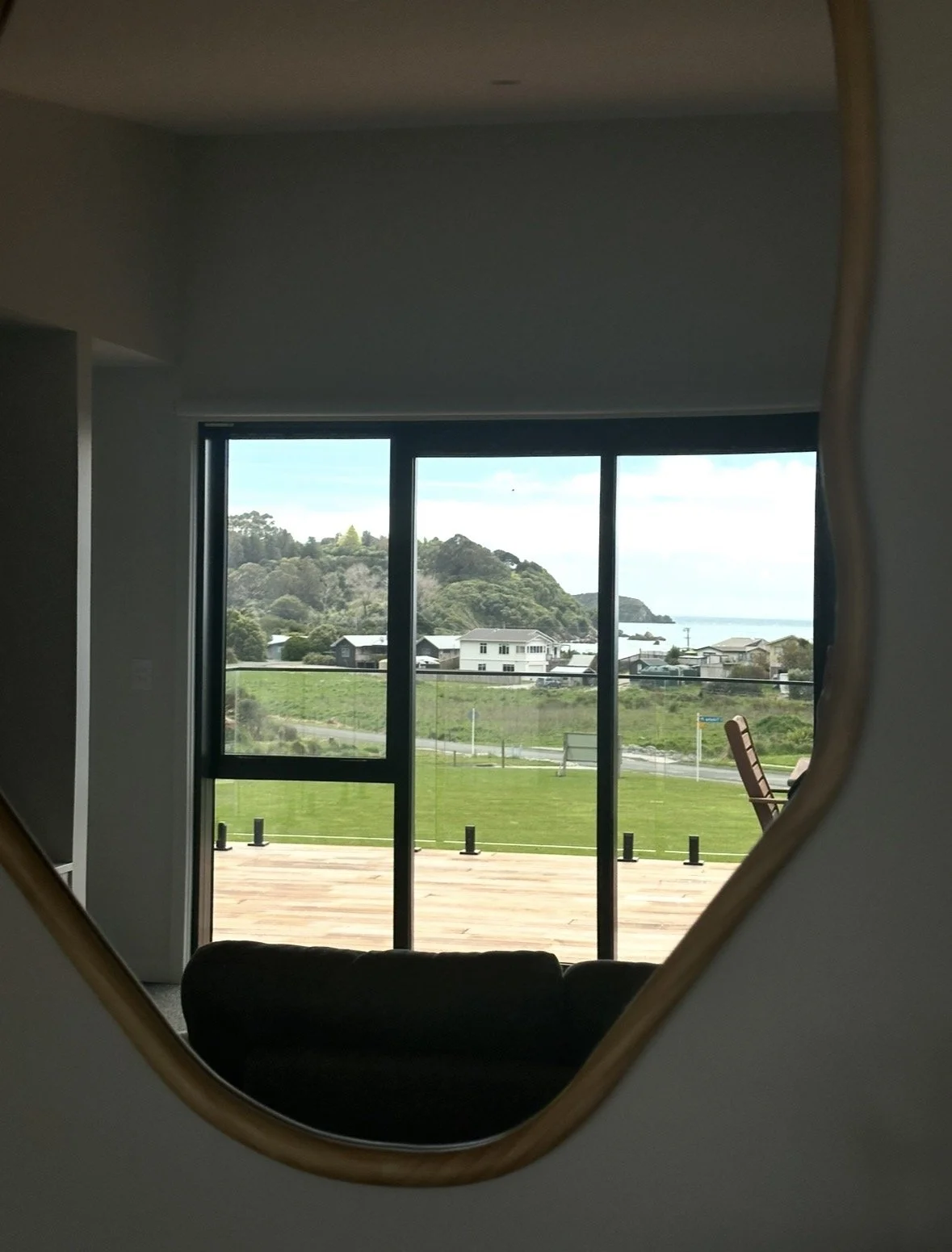Living room view through a wood-framed mirror and floor-to-ceiling windows overlooking a deck, grassy field, coastal village, hills, and ocean