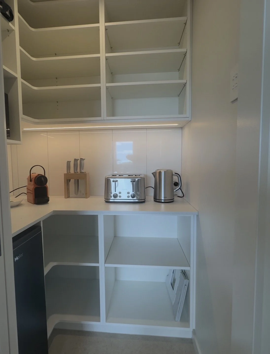 Small white kitchen with open shelving, espresso machine, knife block, toaster, electric kettle, and compact refrigerator