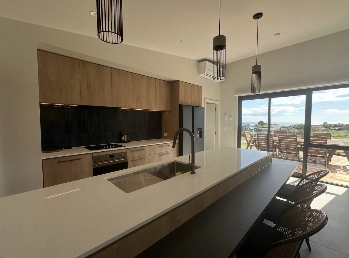 Modern kitchen with wood cabinetry, black backsplash, island sink, pendant lights, bar stools, and sliding doors opening to a sunny patio and ocean view.