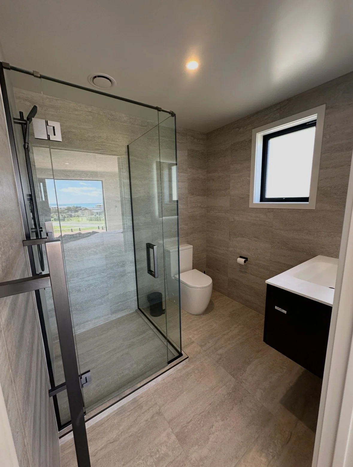 Modern bathroom with a frameless glass walk-in shower, wall-mounted toilet, black vanity, and ocean view