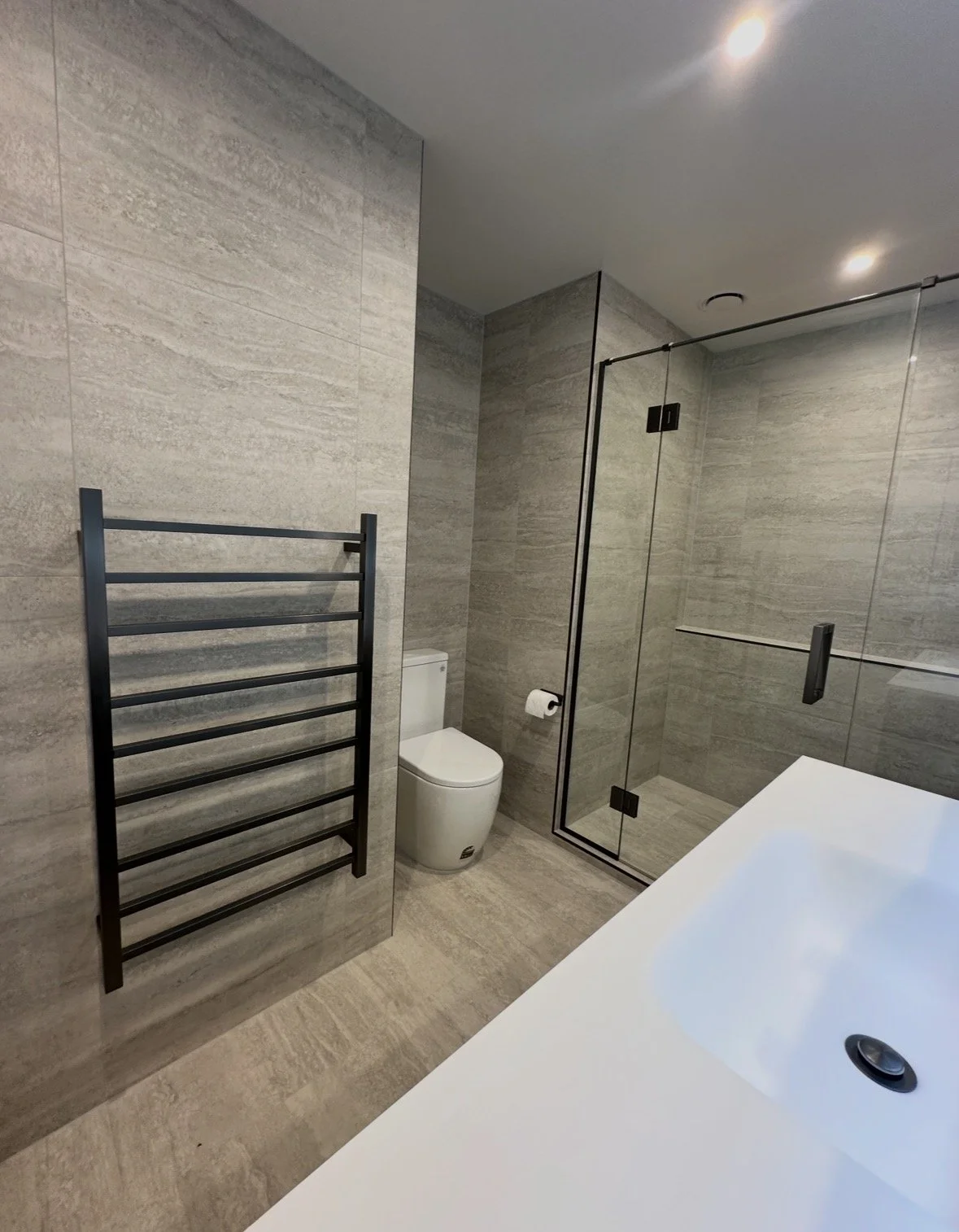 Modern gray-tiled bathroom with a freestanding white bathtub, glass-enclosed shower, toilet, and black heated towel rack