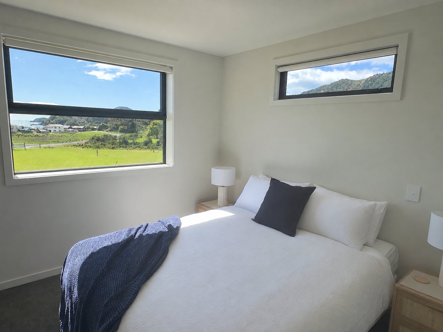 Bright bedroom with a neatly made bed, navy throw blanket, white bedding, bedside tables and lamps, and scenic mountain views through two windows.