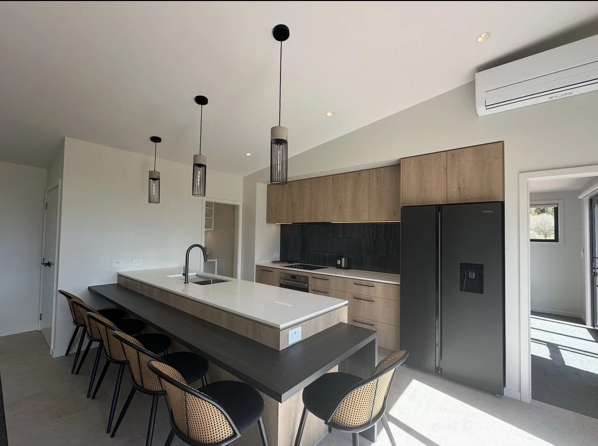 Modern kitchen with wood cabinets, black appliances, island seating, pendant lights, and a white countertop sink.