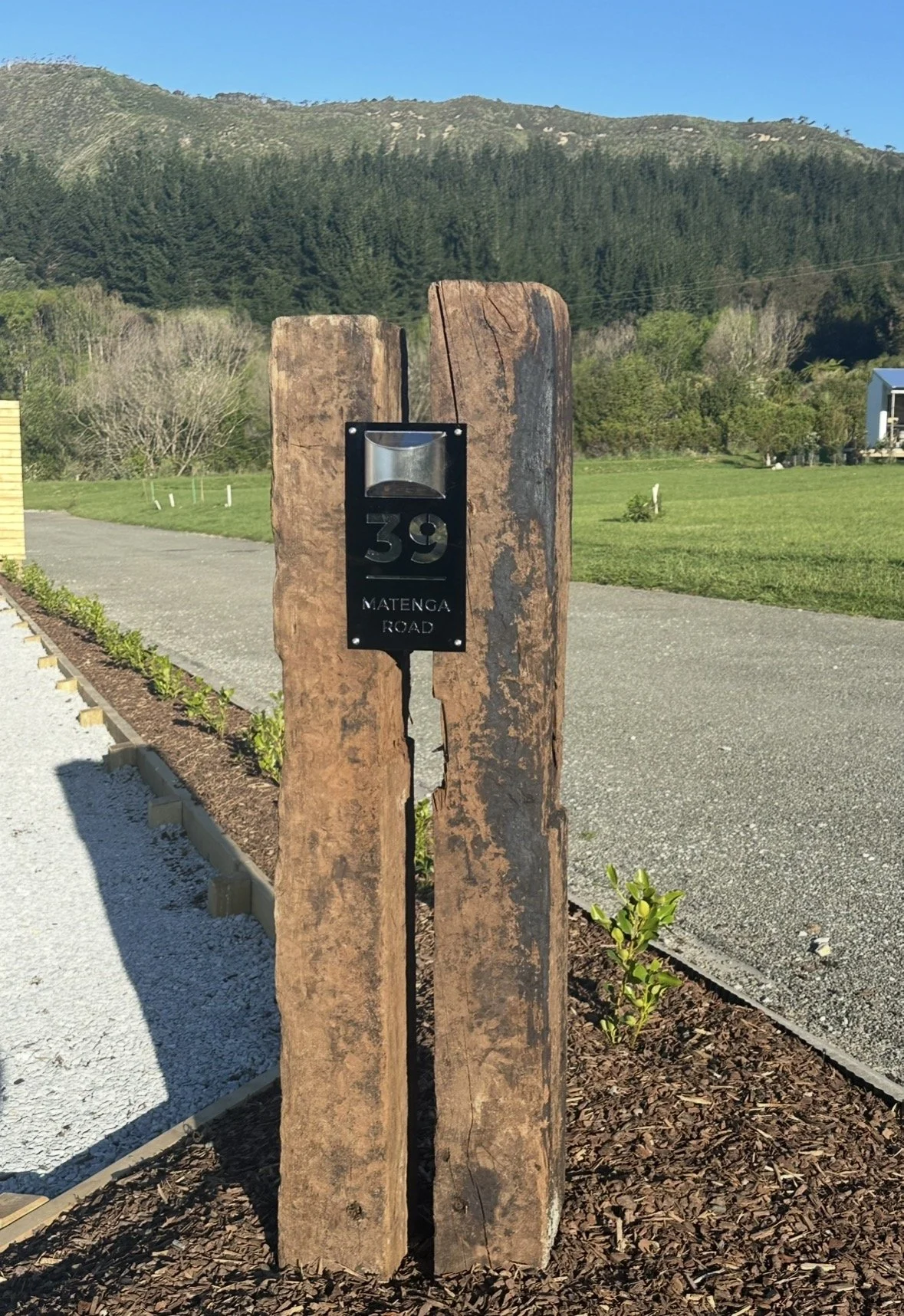 Rustic wooden address marker reading 39 Matenga Road beside a landscaped driveway, with forested hills in the background