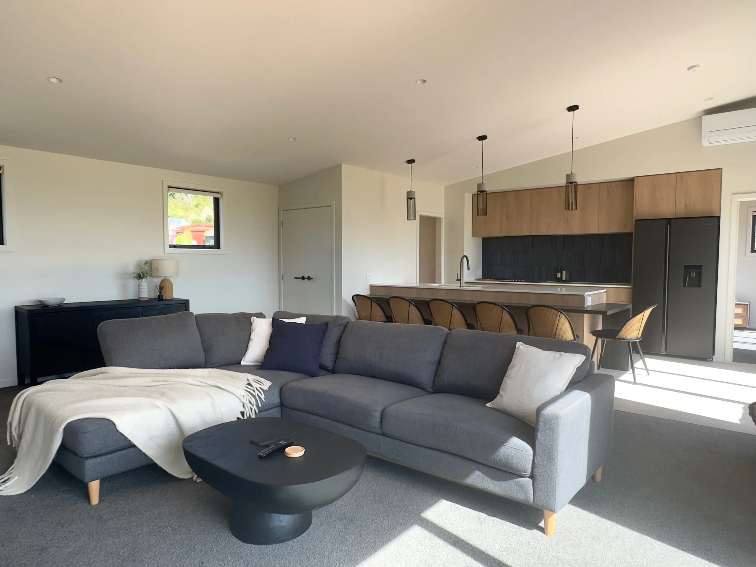 Modern open-concept living room and kitchen with a gray sectional sofa, black coffee table, island seating, wood cabinets, and black refrigerator.