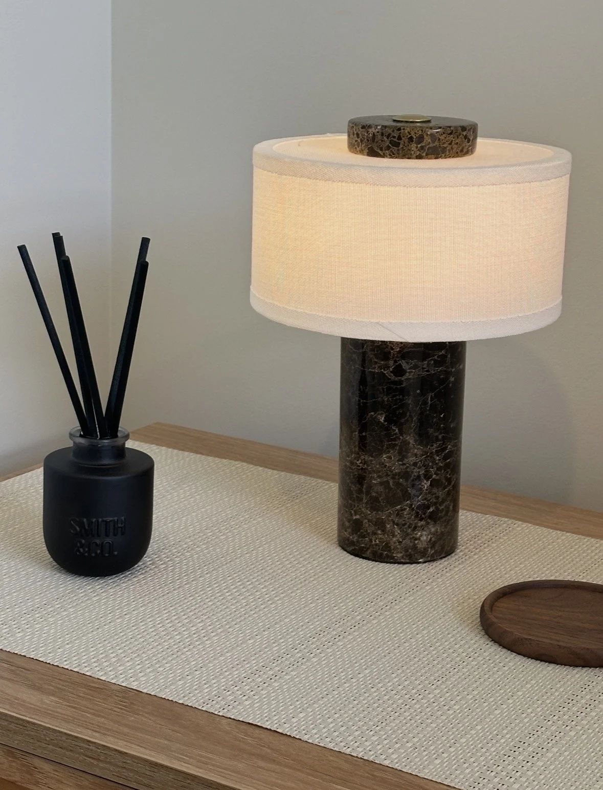 Marble table lamp with a white drum shade beside a black reed diffuser on a wooden console table