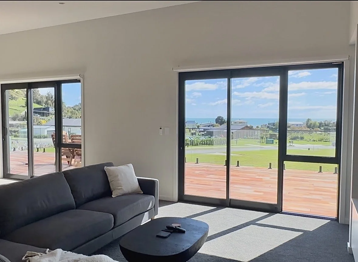 Bright living room with a dark sofa and coffee table overlooking a sunny coastal neighborhood through large sliding glass doors.
