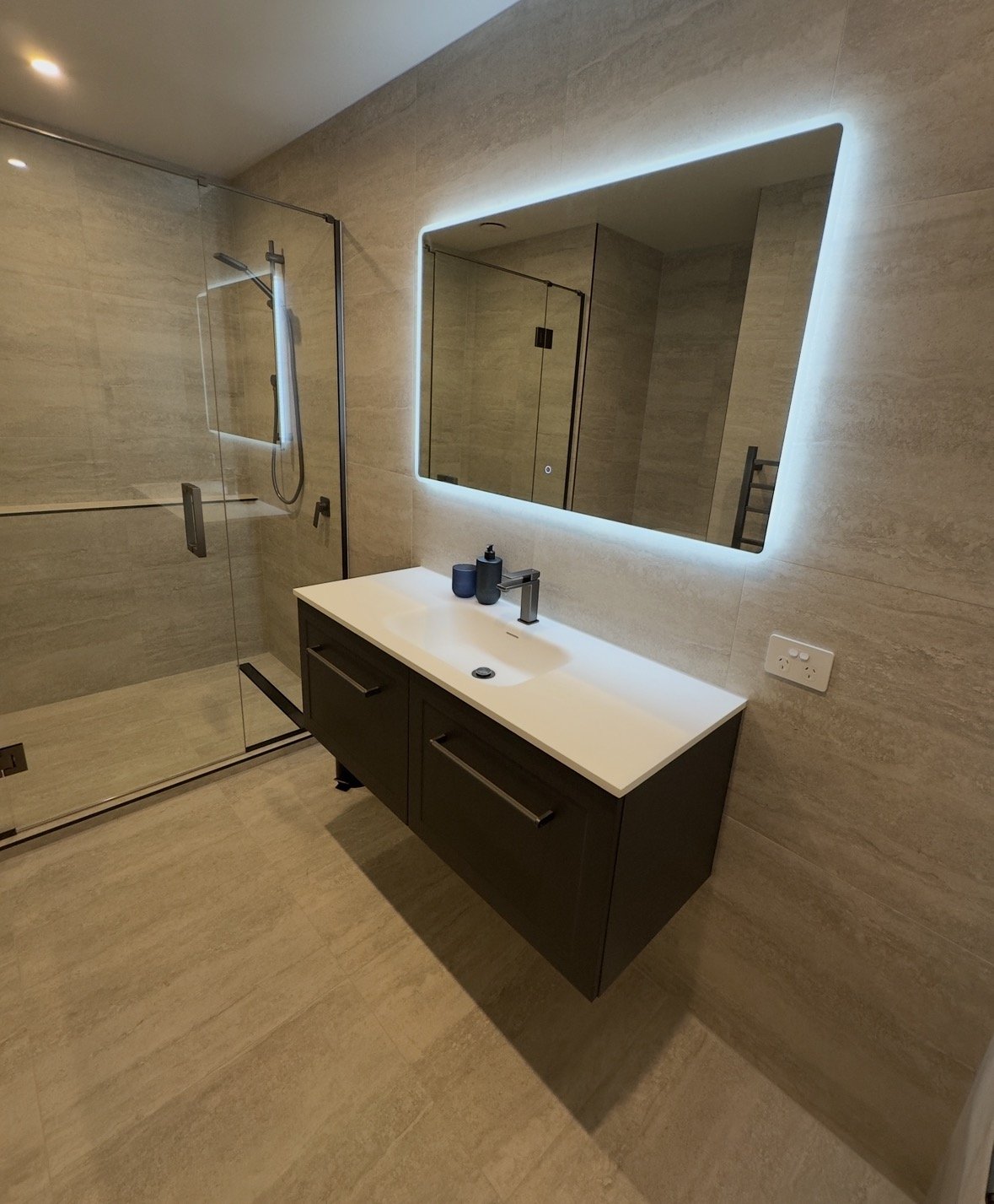 Modern beige-tiled bathroom with a floating dark vanity, white countertop sink, illuminated mirror, and glass-enclosed shower.
