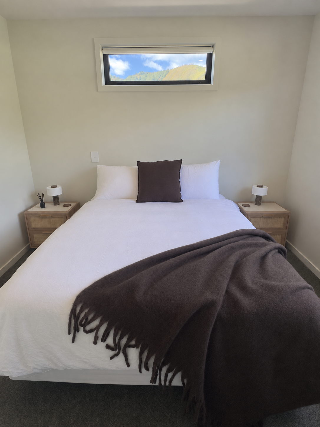 Cozy bedroom with a neatly made bed, dark throw blanket, wooden nightstands, lamps, and mountain views through the window