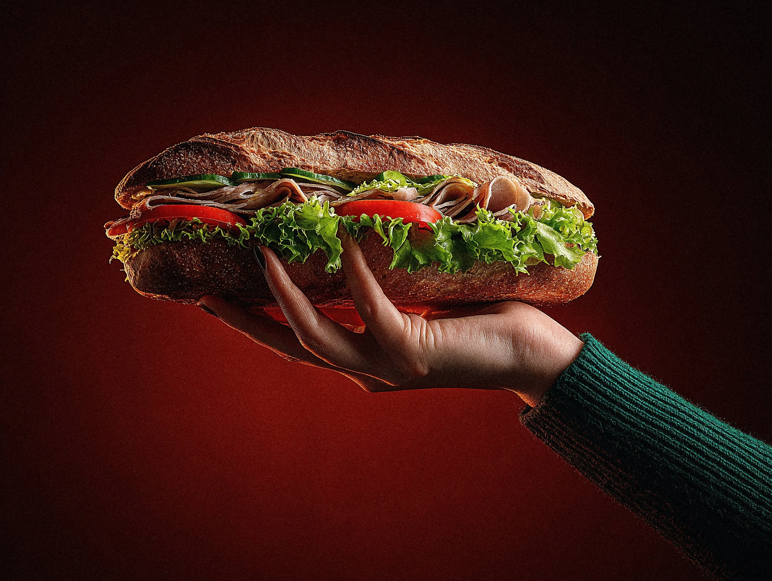 Hand holding a deli sandwich on a crusty baguette with lettuce, tomato, cucumber, and sliced meats
