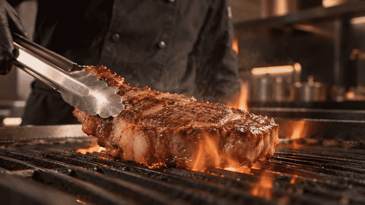 Chef using tongs to grill a seasoned steak over open flames
