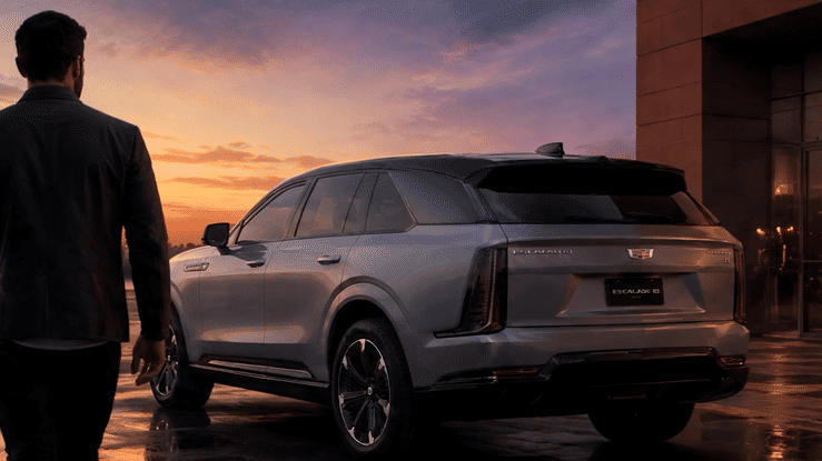 Man standing beside a silver Cadillac Escalade IQ electric SUV at sunset outside a modern building.
