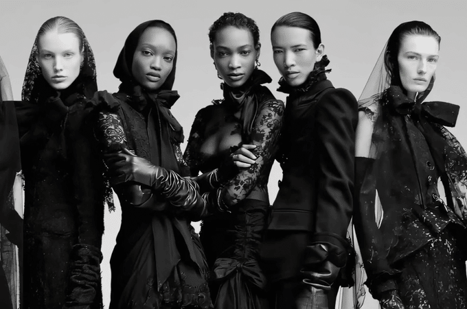 Black-and-white fashion portrait of five models wearing dramatic dark designer clothing and accessories.