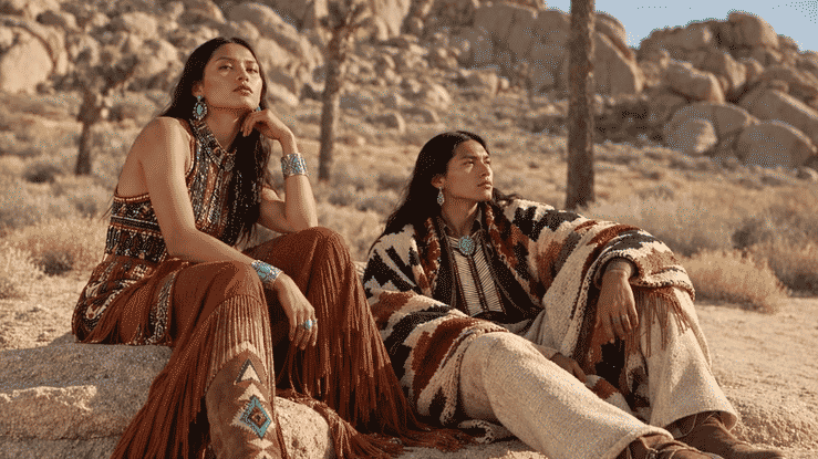 Two Indigenous people in traditional Southwestern clothing seated among desert rocks and Joshua trees.