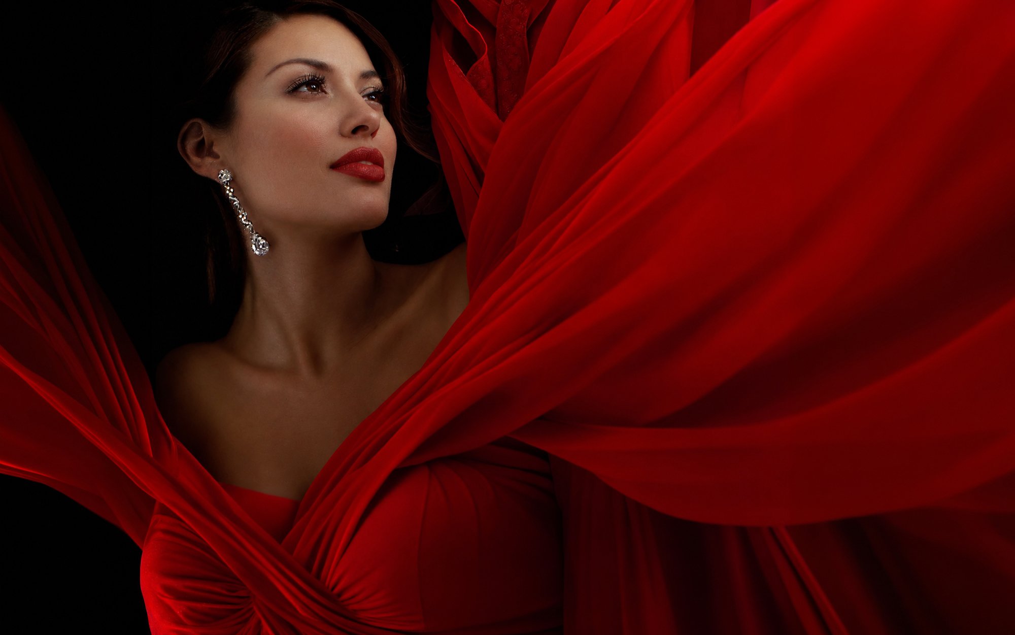 Elegant woman in a flowing red gown with sparkling drop earrings against a black background