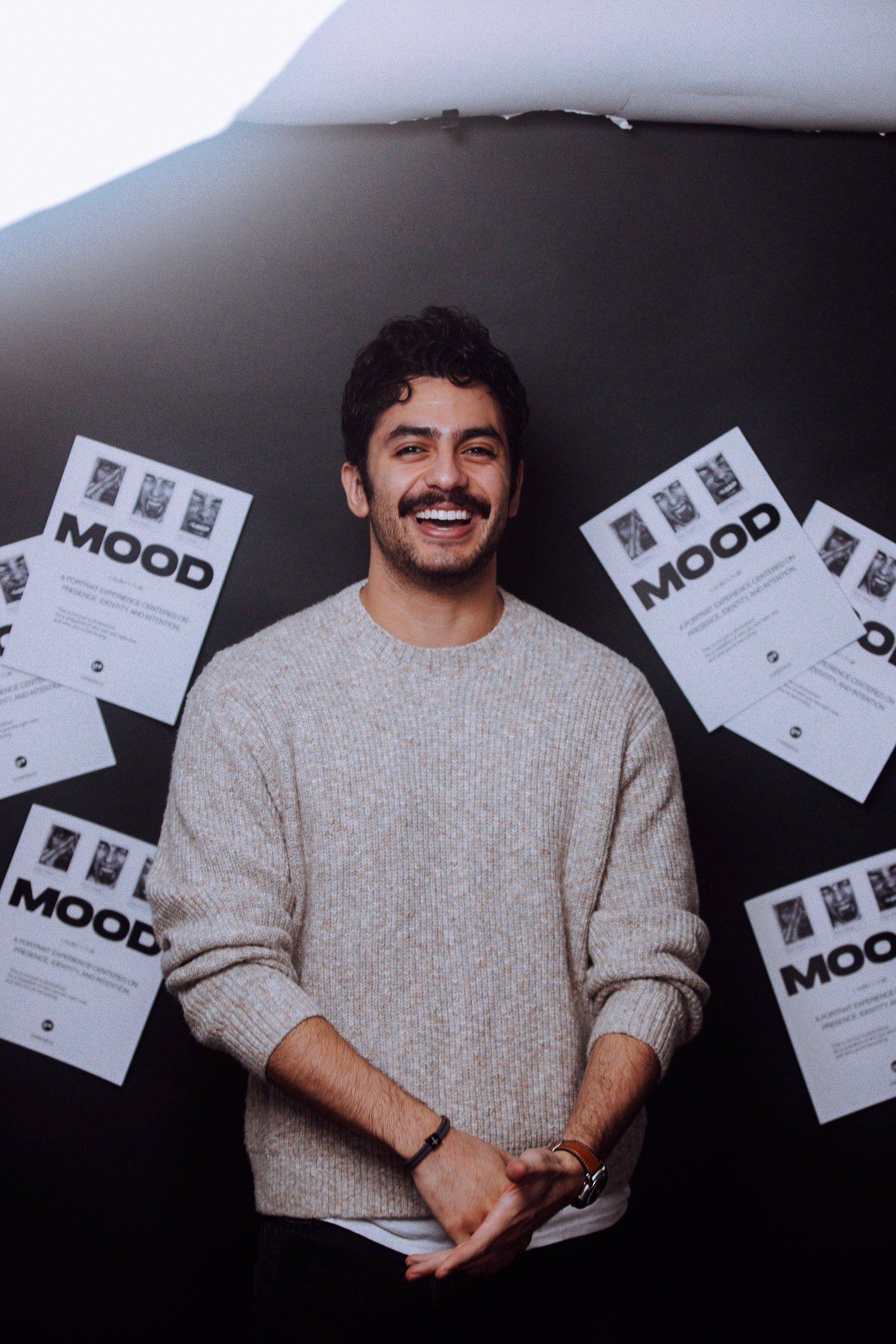 Smiling man in a gray sweater standing in front of posters labeled “MOOD.”