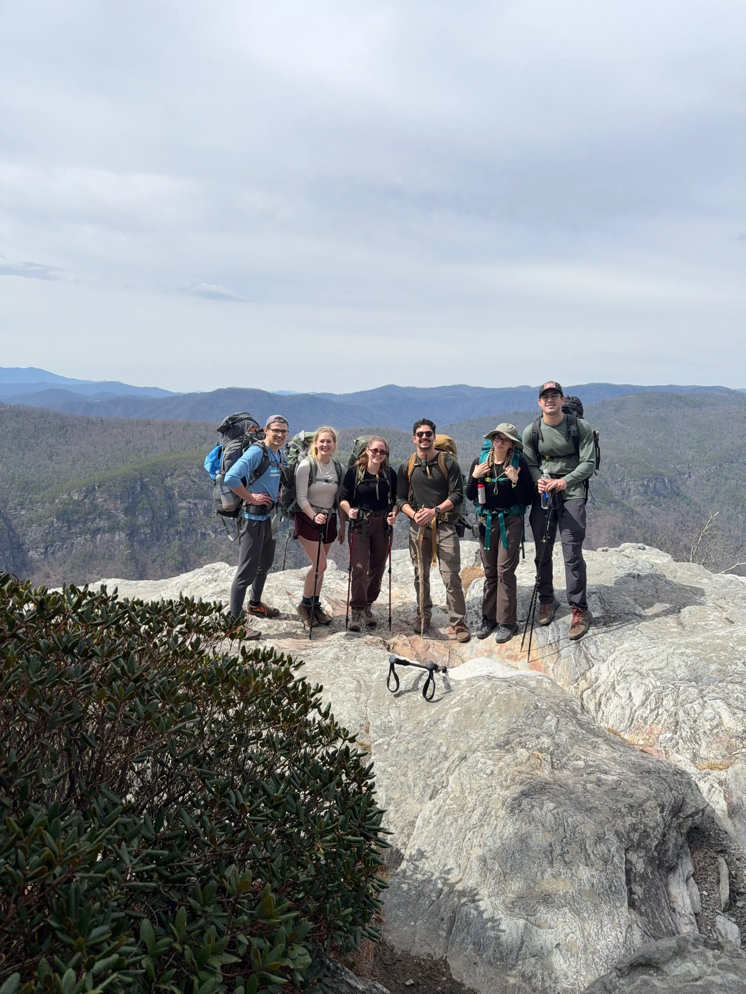 Group of six hikers with backpacks posing on a rocky mountain overlook with forested mountains in the background