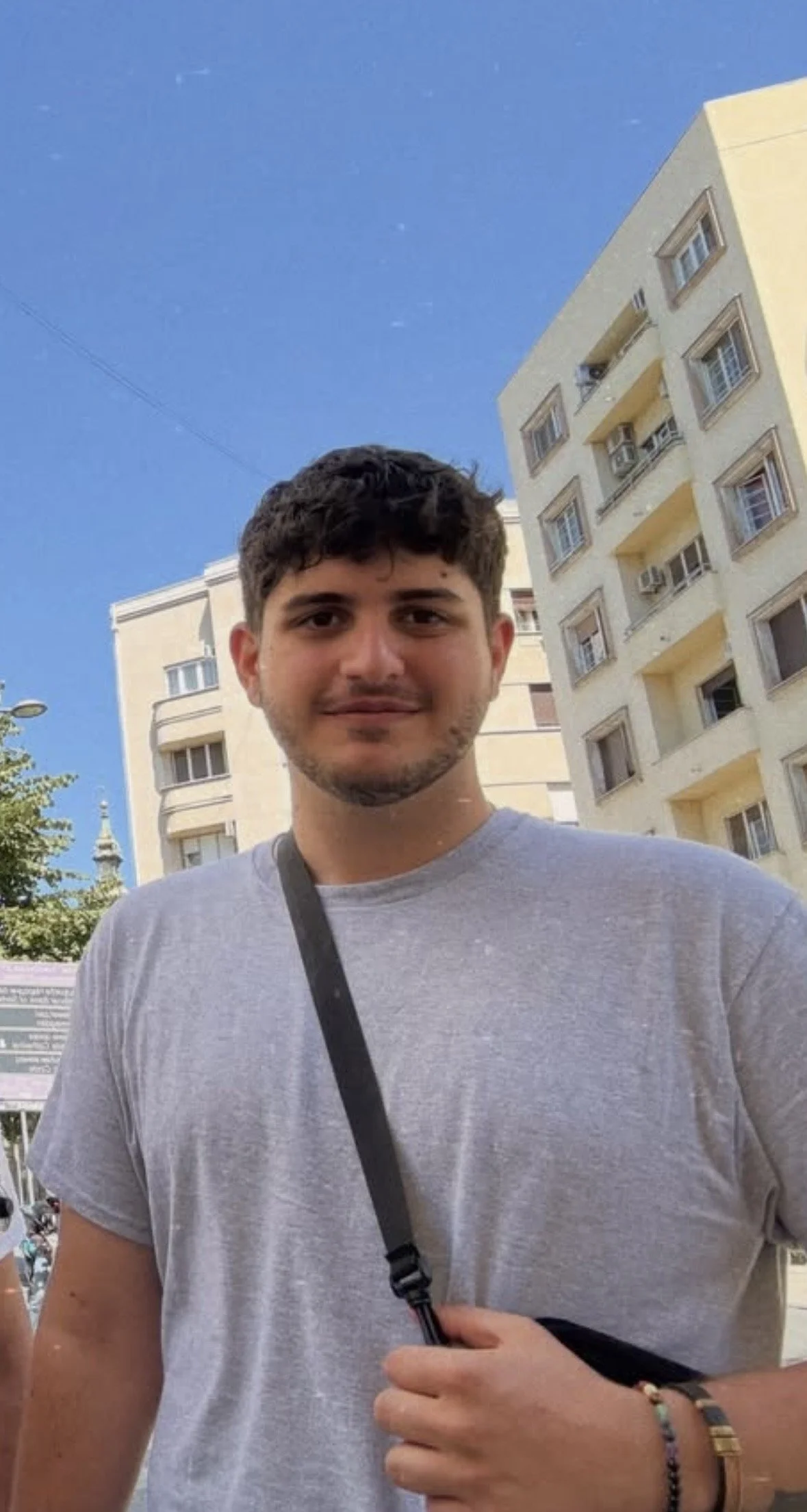 Young man in a gray T-shirt and crossbody bag standing outdoors in front of apartment buildings under a clear blue sky