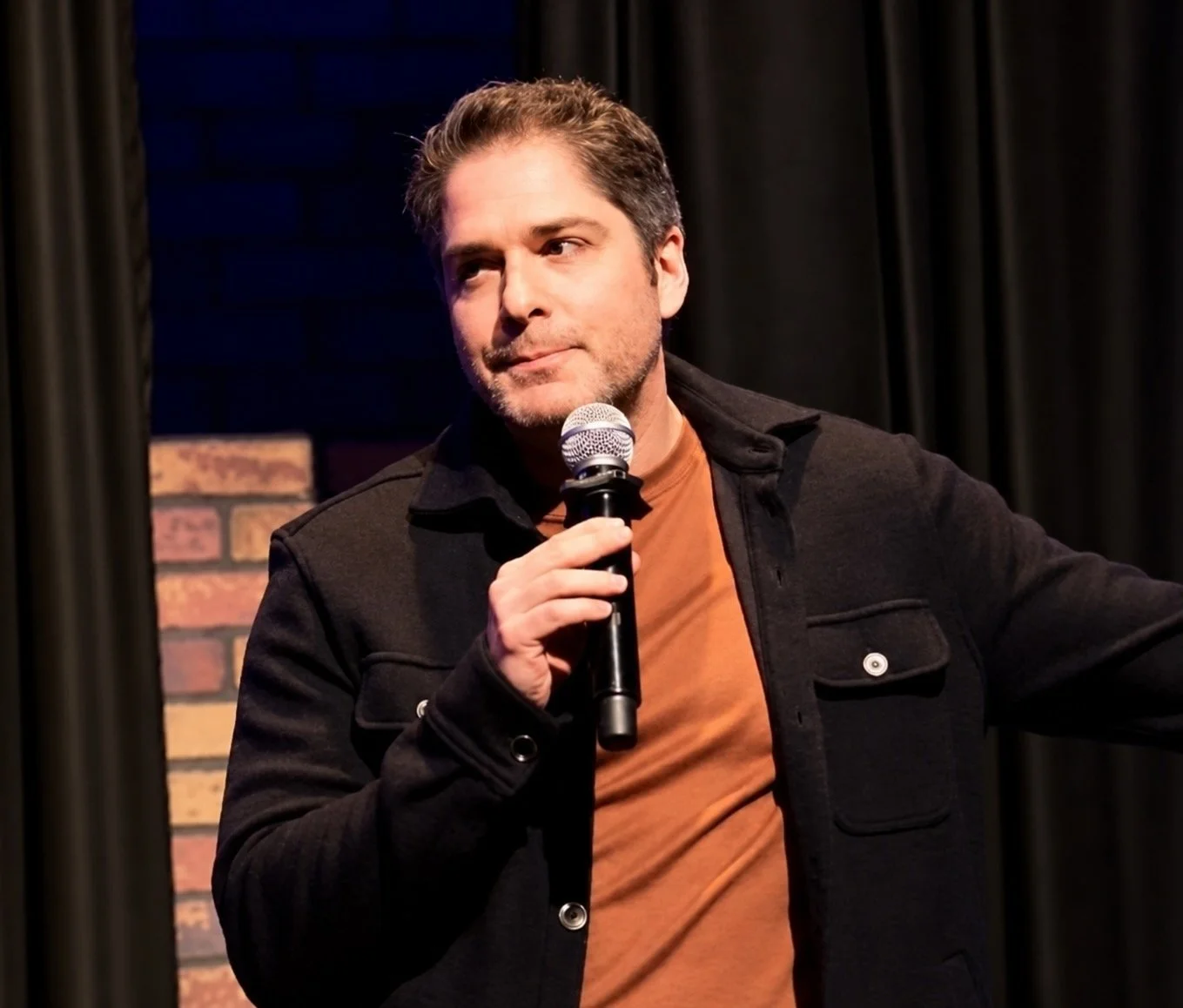 Male comedian holding a microphone on stage in front of a brick wall and black curtains