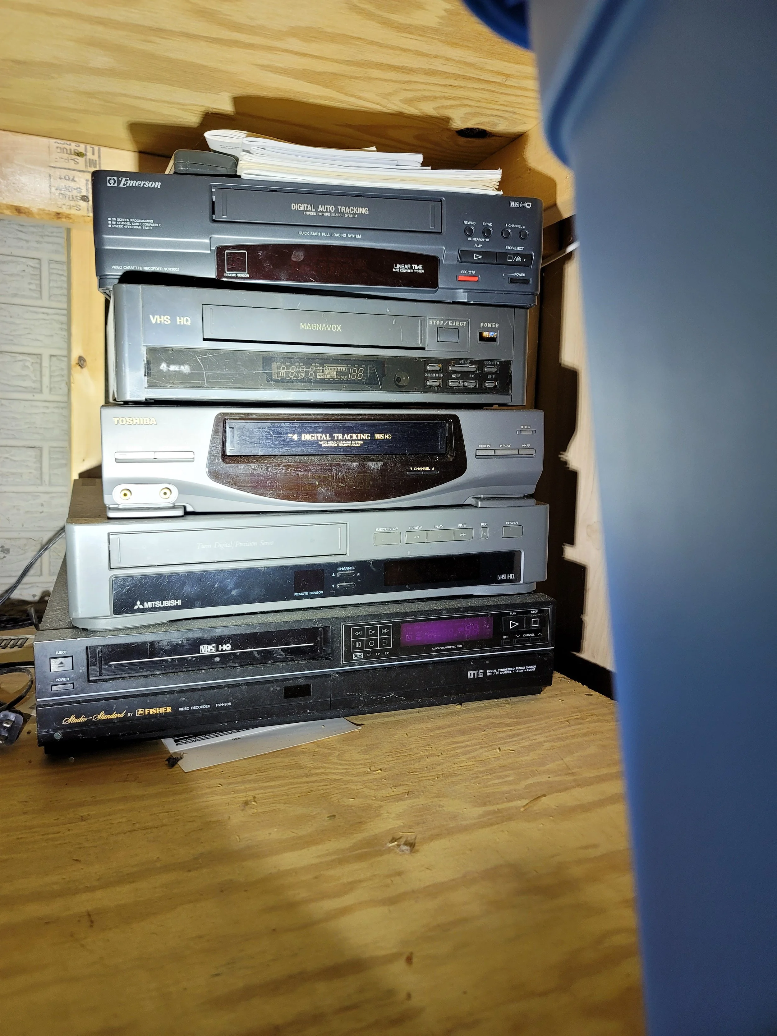 Stack of vintage VHS VCRs from Emerson, Magnavox, Toshiba, Mitsubishi, and Fisher on a wooden shelf