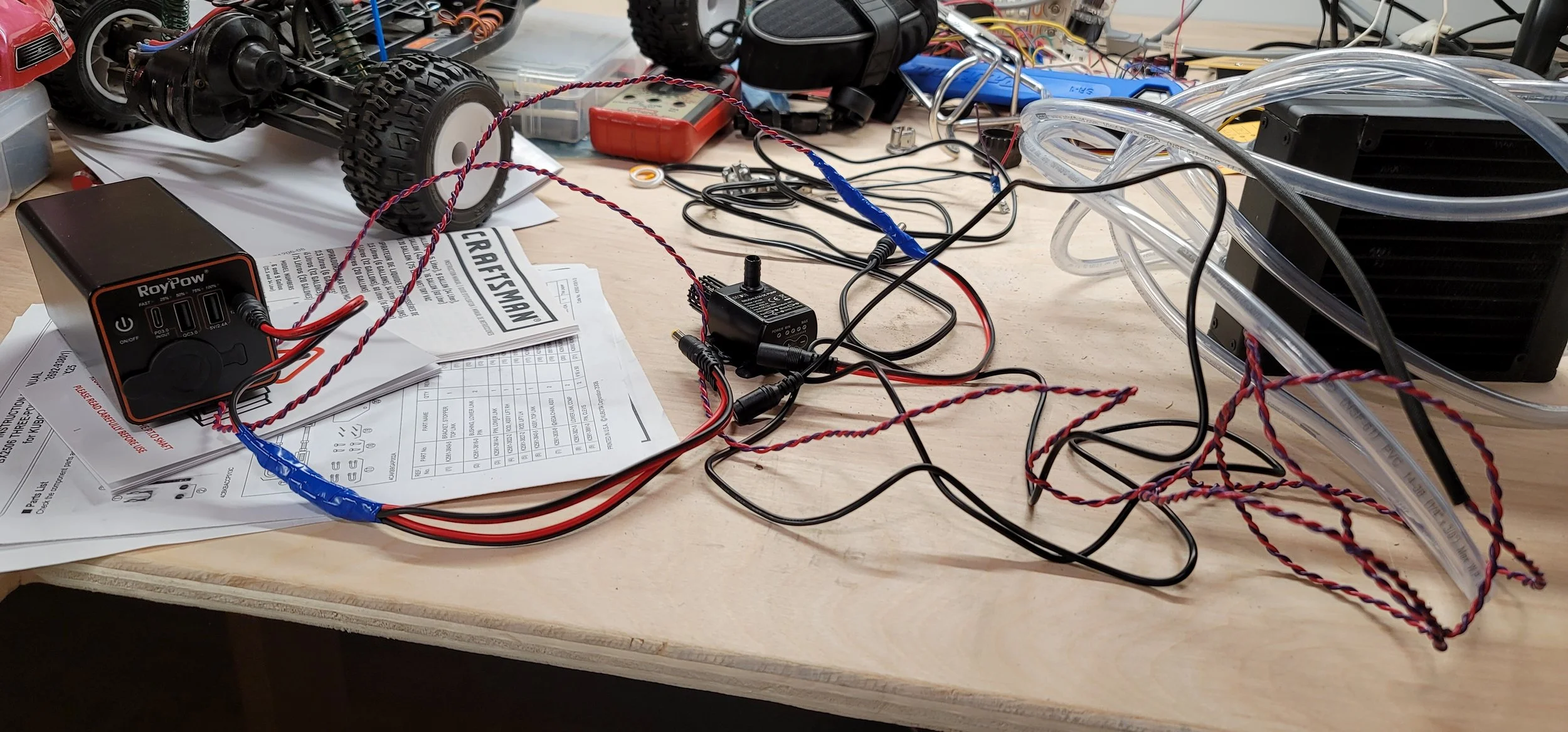 Electronics workbench with an RC off-road vehicle, power supply, pump, tubing, wires, and technical manuals