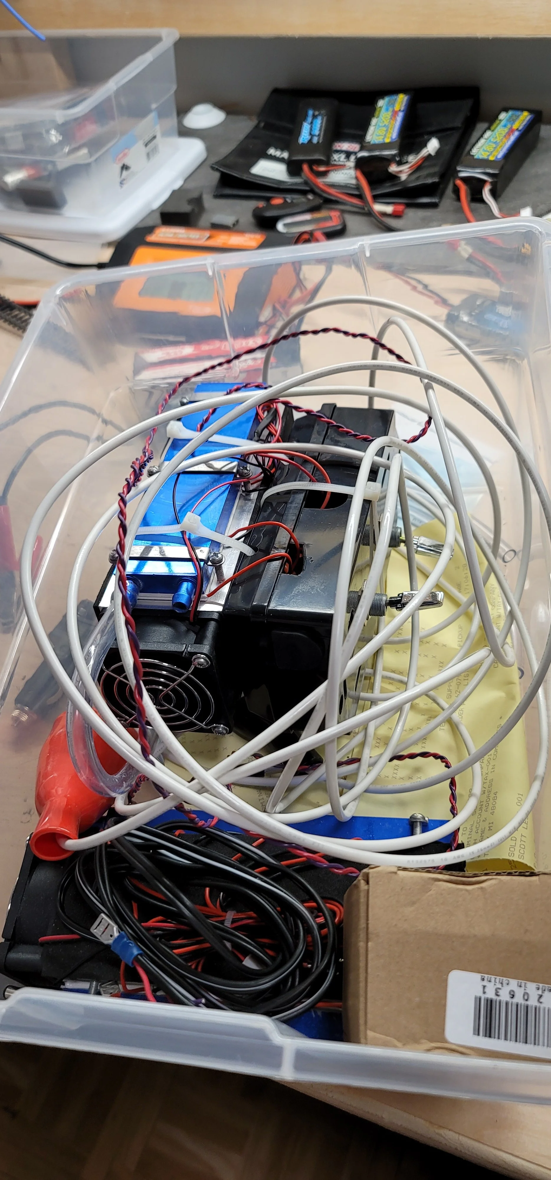 Plastic storage bin filled with batteries, electronic components, circuit boards, and tangled wires on a workbench