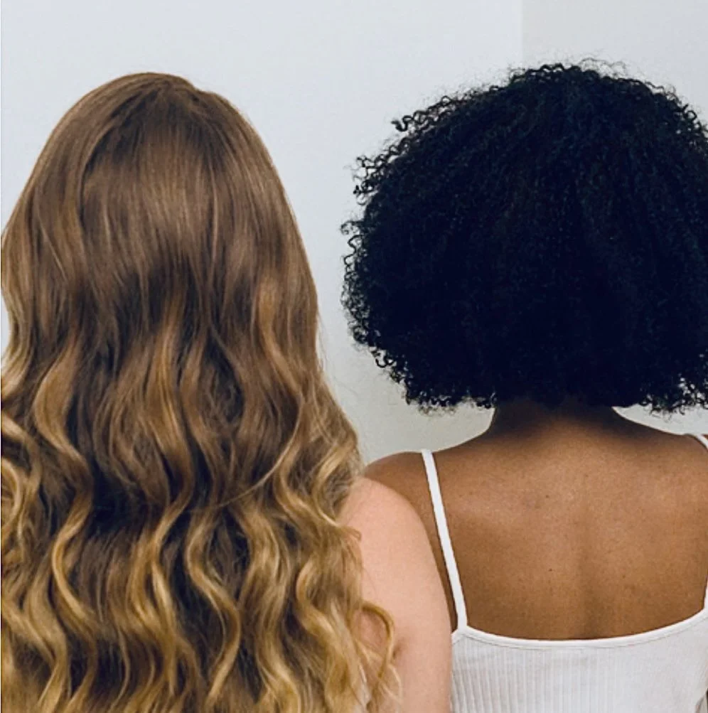 Vue arrière de deux femmes aux textures de cheveux différentes : l’une aux longs cheveux ondulés châtains, l’autre aux cheveux afro courts et bouclés.