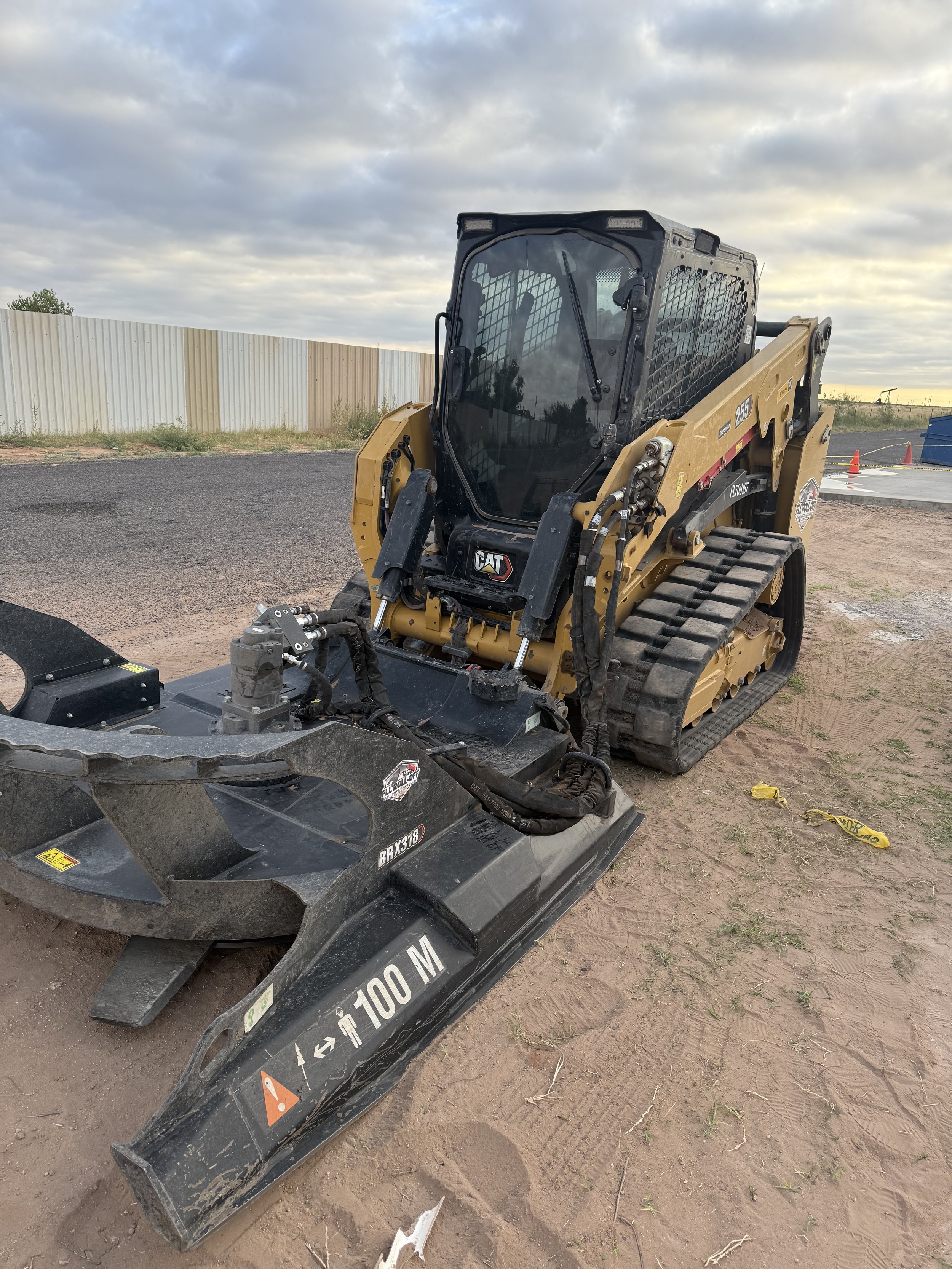 Yellow CAT tracked skid steer loader with a large black attachment parked at a construction site under a cloudy sky.
