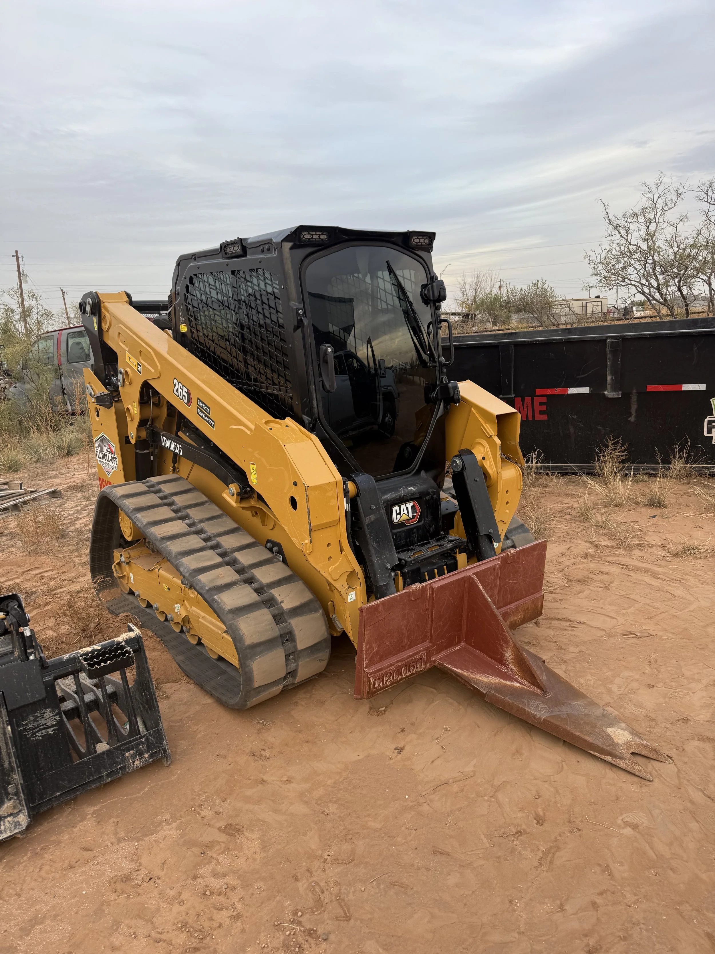 Yellow Caterpillar tracked skid steer loader with a front bucket parked on a dirt lot beside a black dump trailer.