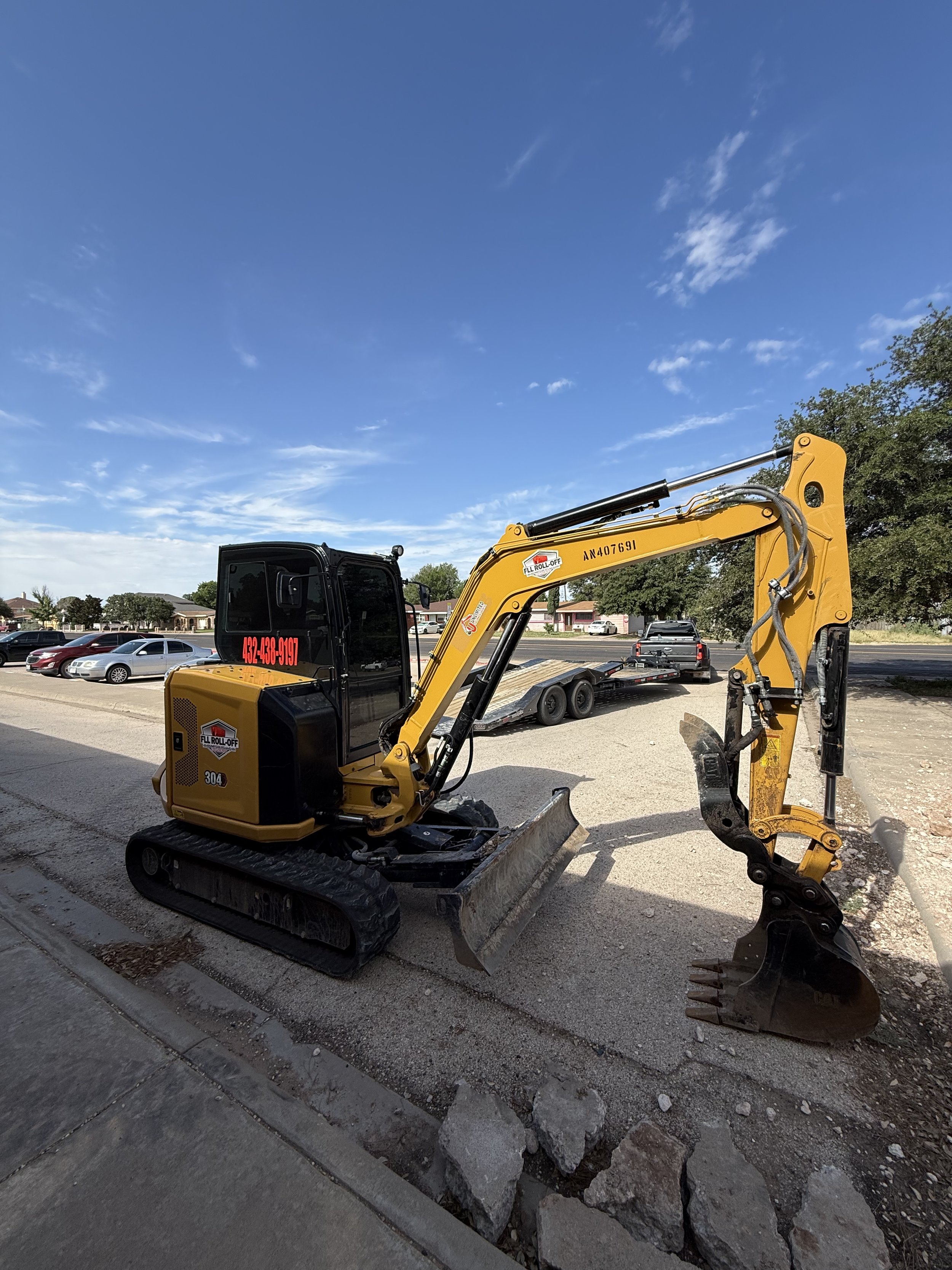 Yellow tracked excavator with hydraulic arm and bucket parked beside a street, trailer, and passing cars under a blue sky