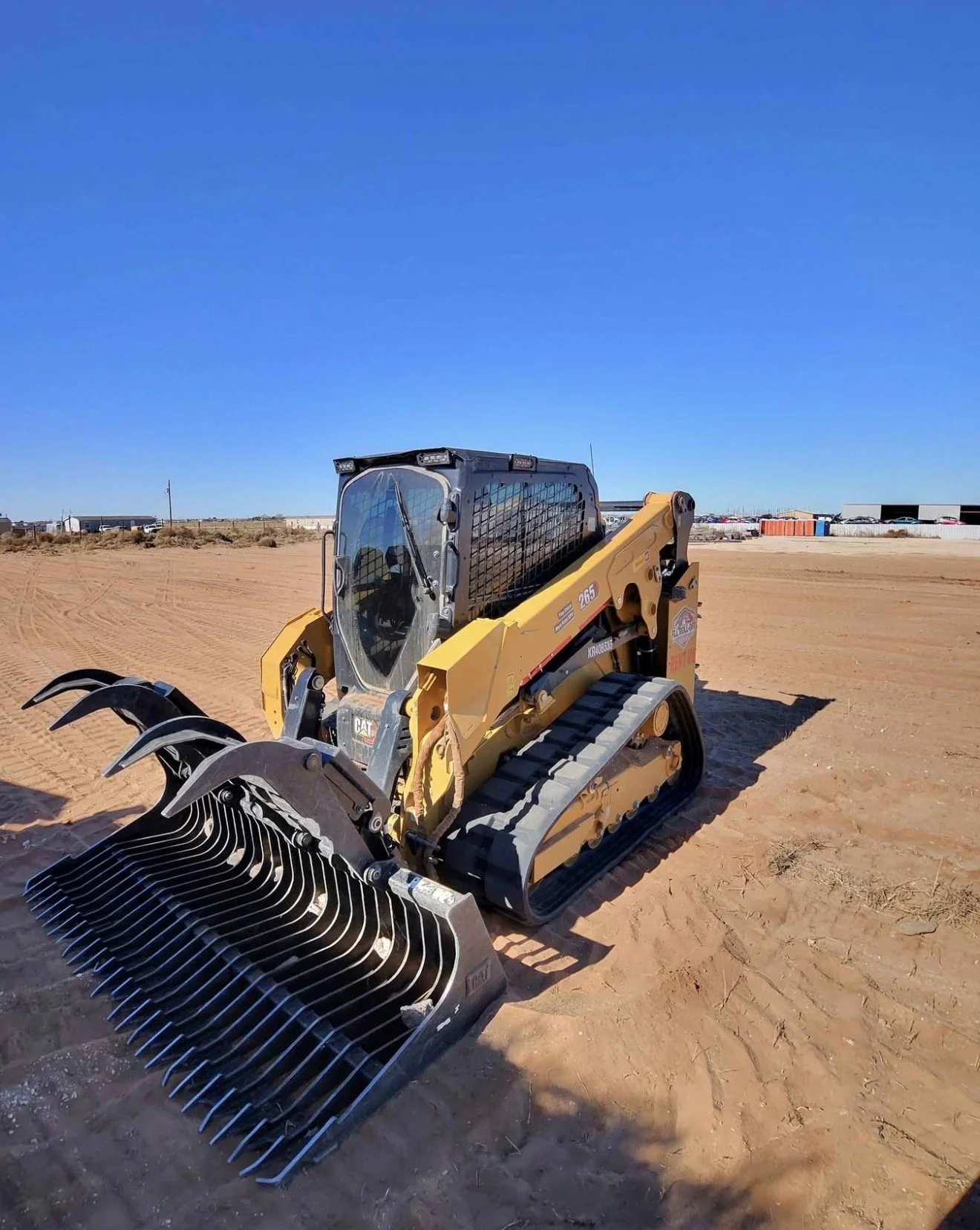 Yellow CAT compact track loader with a grapple attachment parked on sandy ground at an outdoor equipment yard.