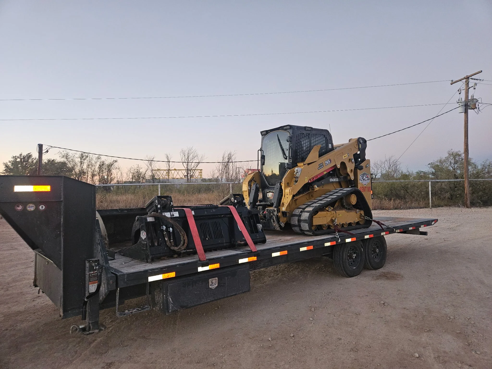 Caterpillar tracked skid steer loader and attachments secured on a flatbed utility trailer at dusk