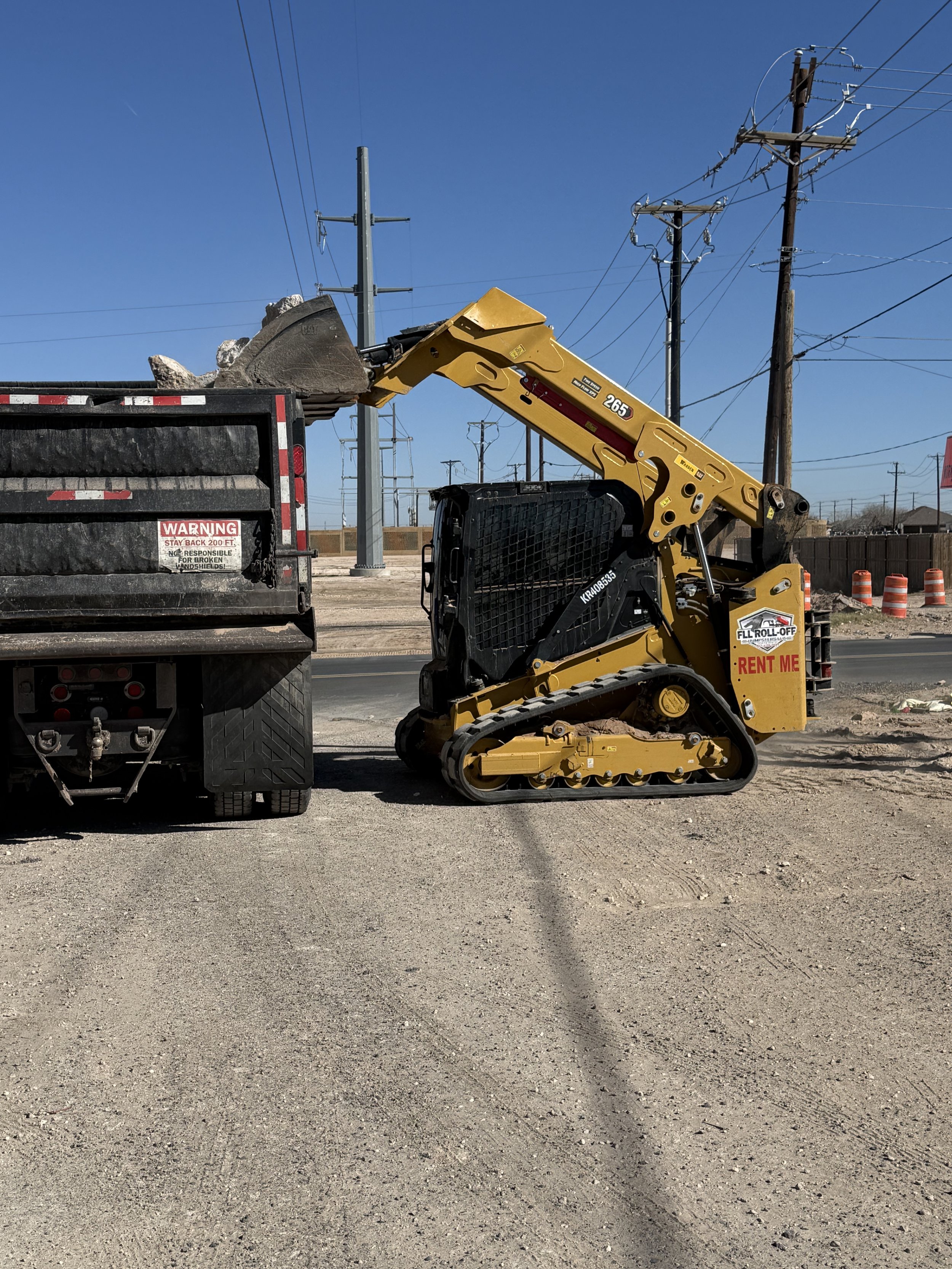 Yellow tracked skid-steer loader with a hydraulic breaker attachment beside a dump truck at a construction site.
