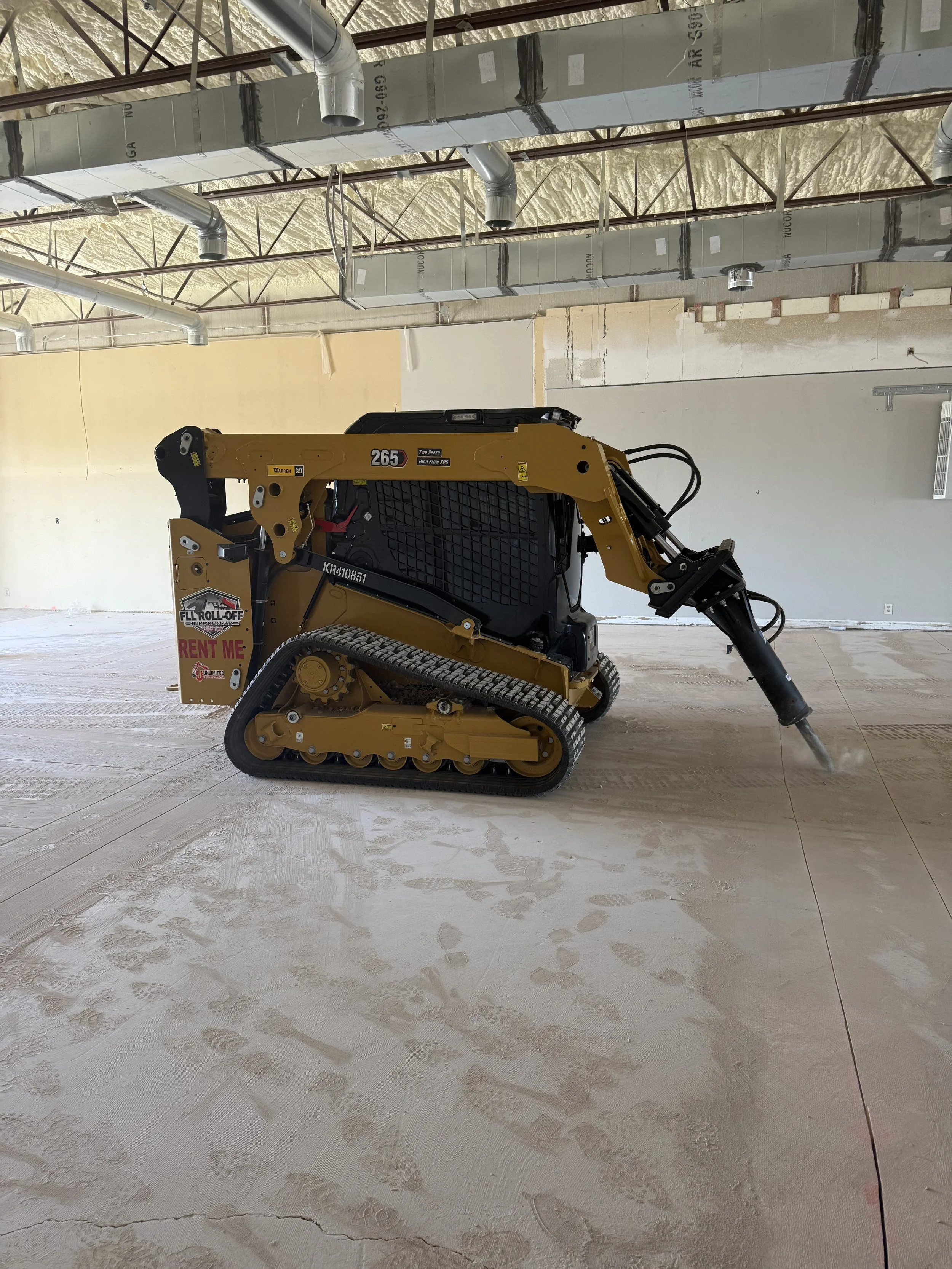 Yellow tracked Caterpillar 265 skid steer with hydraulic attachment inside a partially renovated building.