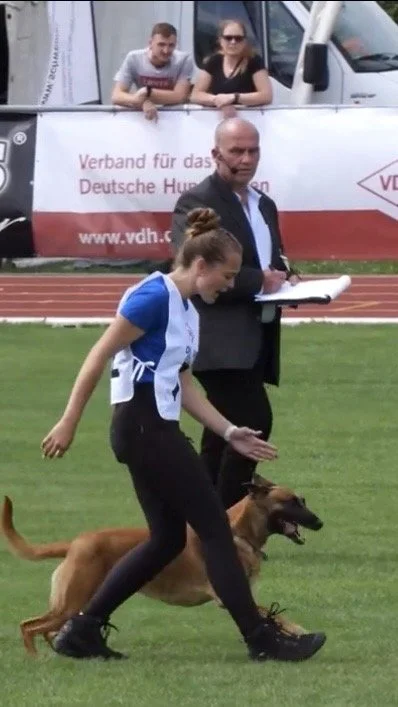 Eine junge Frau trainiert mit einem Hund auf einer Wiese, während ein Mann in Anzug mit Notizblock zuschaut. Im Hintergrund beobachten zwei Personen die Szene von einer Absperrung.